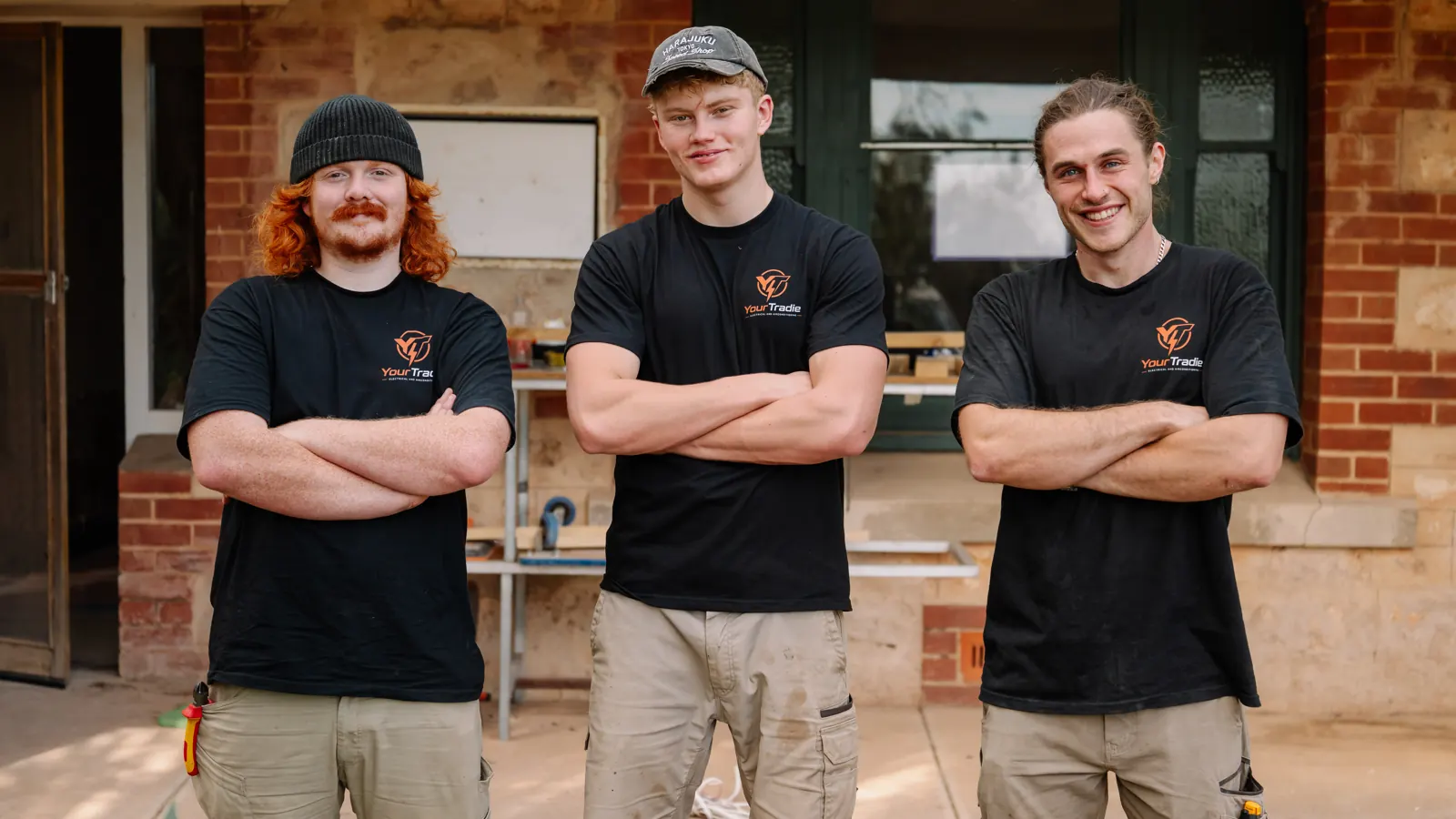 The YourTradie crew, three tradies in branded black work shirts with arms crossed outside a stone villa
